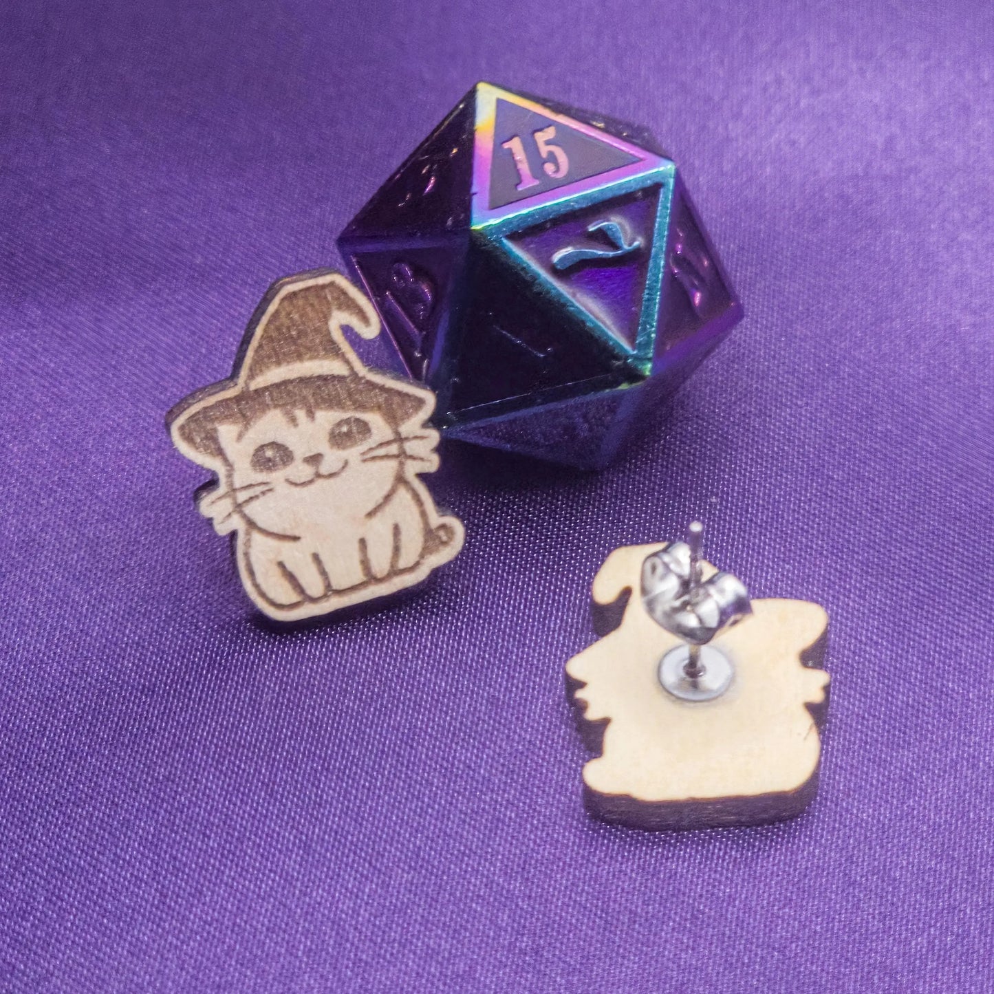 Wooden cat-shaped earrings with a witch hat design and a metallic die on a purple background.