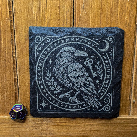 Engraved slate tile featuring a detailed raven holding a key in its beak, surrounded by runes and crescent moon, next to a colorful 12-sided dice.