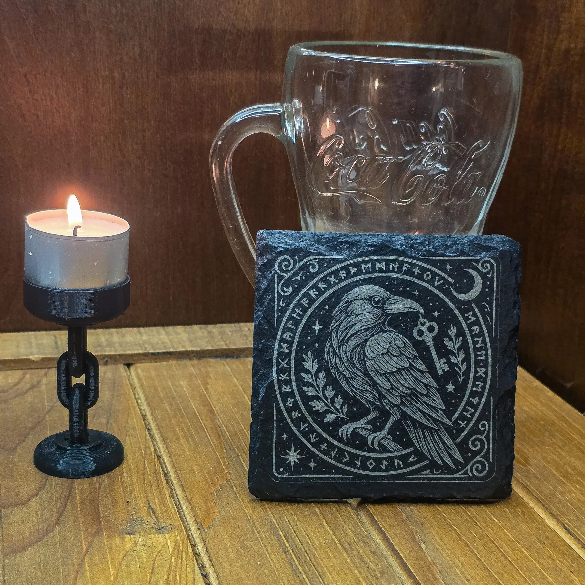 A lit candle in a black holder sits on a wooden table next to a glass mug and a black stone coaster with an intricate raven and rune design. Moody and mystical atmosphere.