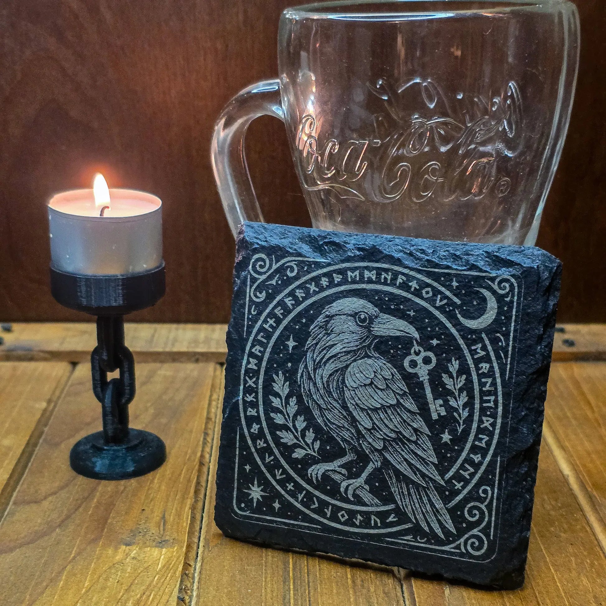 A lit candle on a stand, a clear mug with cursive lettering, and a slate with a detailed etching of a raven holding a key, surrounded by runes. Cozy, mystical ambiance.