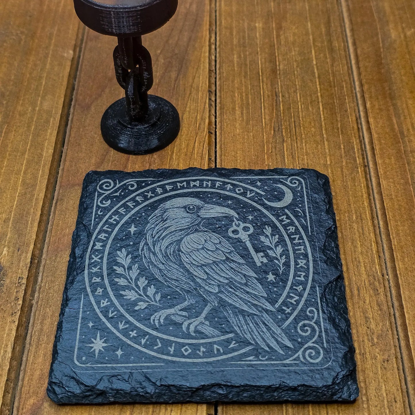 A black slate coaster with an engraved raven holding a key is placed on a wooden table. The coaster features ornate designs and runic symbols, creating a mysterious, mystical tone.