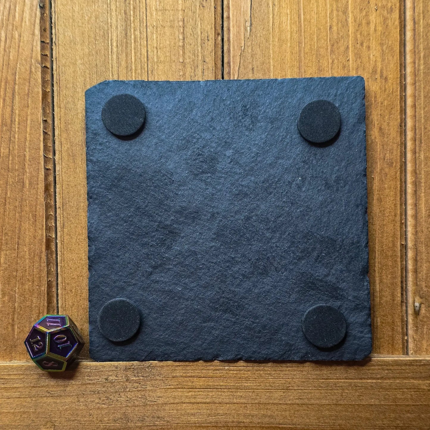 A black slate coaster with round, black felt pads on each corner is placed on a wooden surface. A multicolored twenty-sided die is positioned beside it.