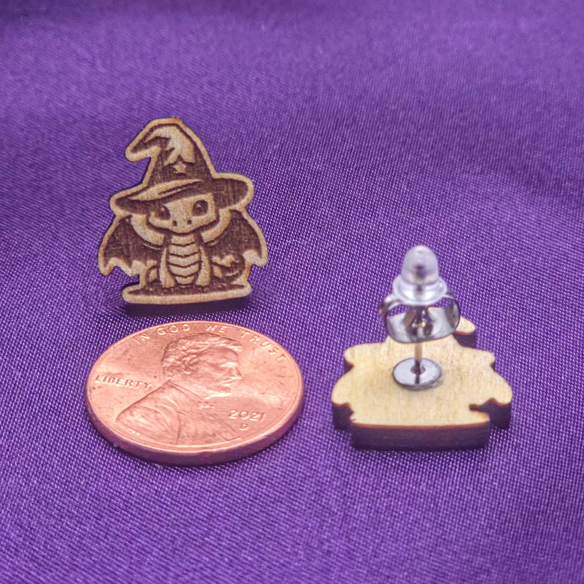 Two small wooden laser engraved earrings featuring cute dragons with wizard hats on a purple surface with a penny for scale.