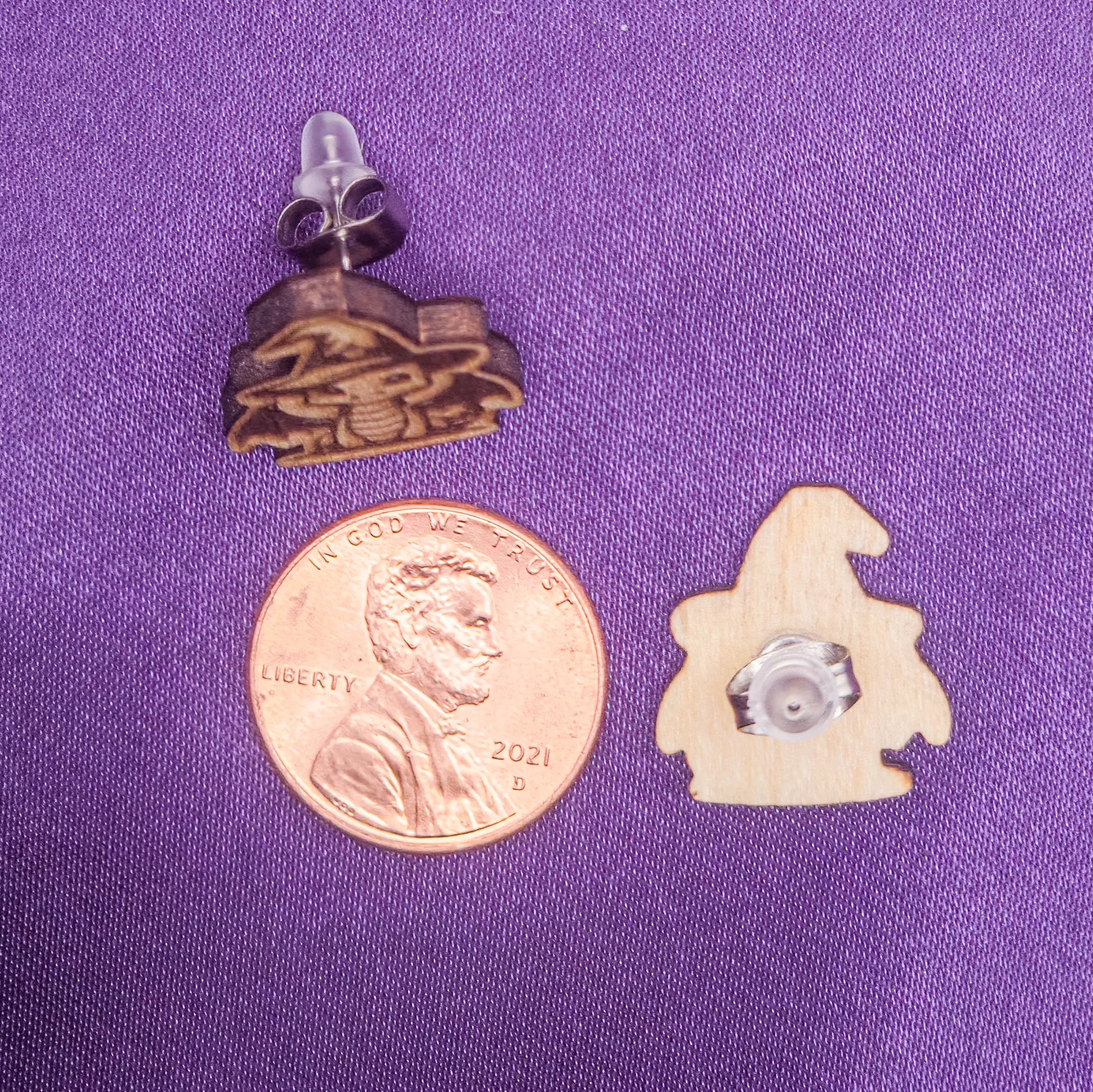 Two small wooden laser engraved earrings on a purple surface with a penny for scale.