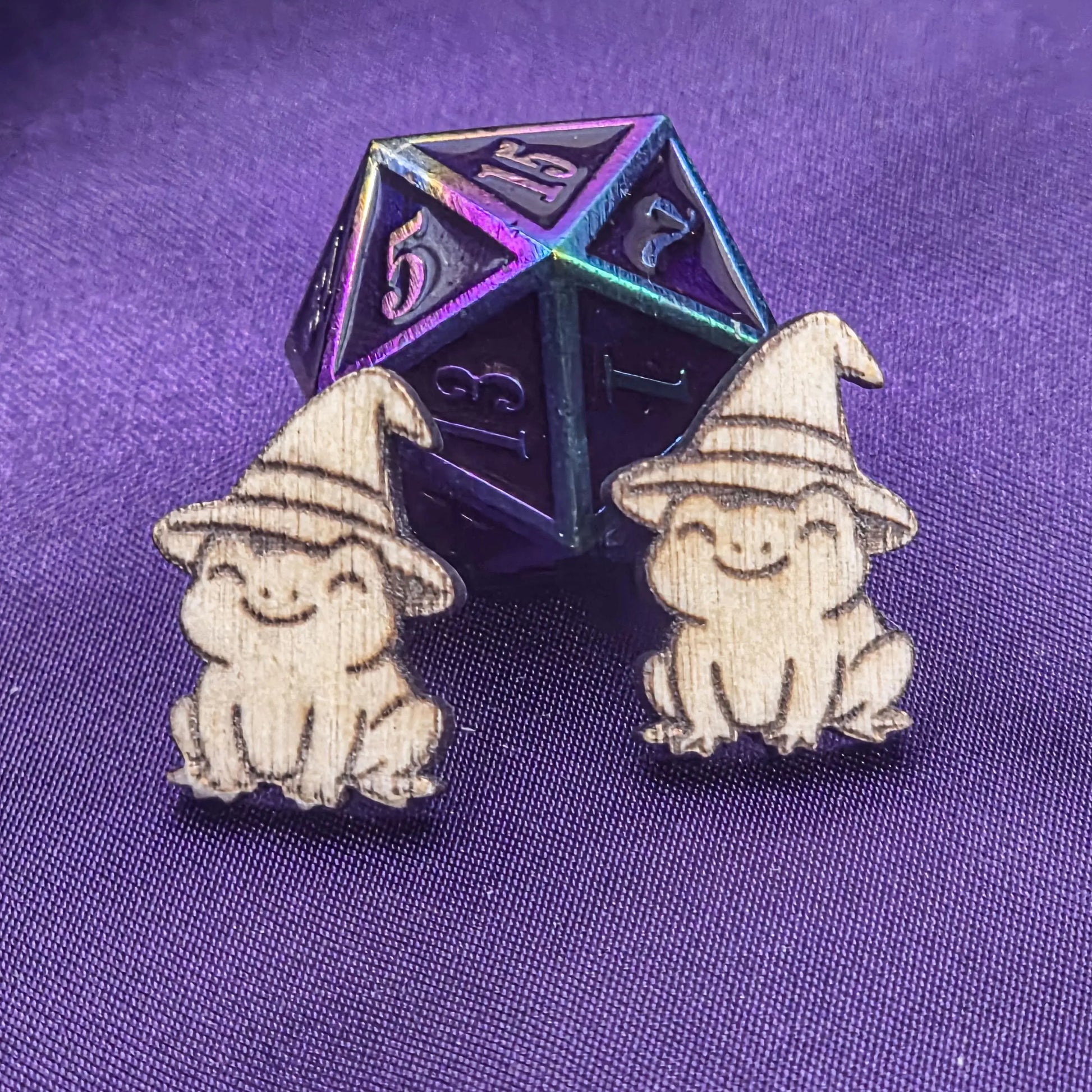 Two small laser engravings featuring frogs wearing hats on a purple background with a metallic polyhedral die.