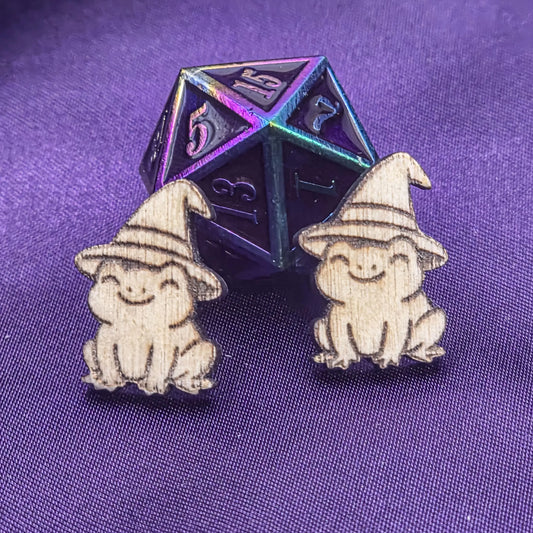 Two small laser engravings featuring frogs wearing hats on a purple background with a metallic polyhedral die.